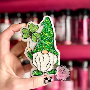 May include: A St. Patrick's Day gnome-shaped decoration holding a shamrock. The gnome has a white beard, a green glitter hat, and green glitter accents. The decoration is held in a hand.