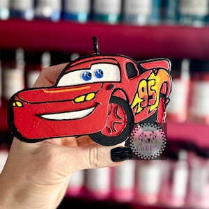May include: Red Lightning McQueen car-shaped ornament with black outlines and a loop for hanging. The car features the number 95 in yellow and orange, blue eyes, and a smiling mouth. The ornament is held in a hand.