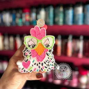 May include: A white, pink, green, and black glittery chicken charm with green glasses. The charm has a pink beak and black eyes. The charm is held in a hand.