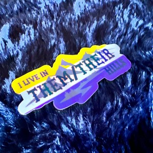 May include: A sticker with a yellow, white, and purple color scheme. The sticker features a mountain range with the text "I live in them/their hills" in black.