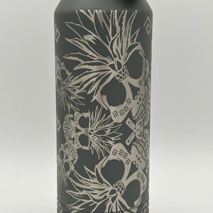 May include: A dark grey water bottle with a black handle and lid. The bottle has an engraved design of skulls and floral elements. The design is in a lighter grey colour, creating a contrast against the bottle's surface.