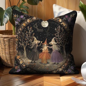 May include: Square decorative pillow featuring a whimsical illustration of three witches dancing around a bonfire under a full moon. The design is set within a floral border on a black background. The witches wear long dresses and pointed hats.