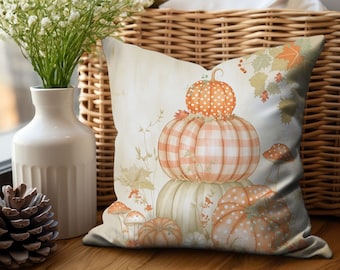 May include: A square throw pillow featuring a printed illustration of stacked pumpkins in shades of orange, white, and muted green. The design includes polka dot and gingham patterns, small mushrooms, and autumn leaves on a light, cream-colored background.