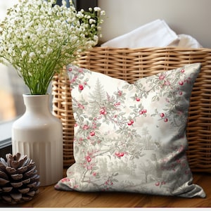 May include: Decorative throw pillow with a vintage-style landscape design. The pillow features a cream-colored background with a pattern of red berries, green leaves, and a scenic landscape in gray tones. The pillow is placed on a wooden surface.