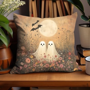 May include: Square decorative pillow featuring a whimsical Halloween scene. Two ghost figures stand in a field of colorful flowers under a full moon, with bats flying above. The pillow has a vintage, painterly style.