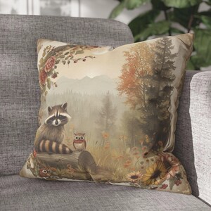May include: A throw pillow with a woodland scene featuring a raccoon, an owl, and sunflowers. The background is a misty forest with a log in the foreground.