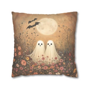 May include: Square throw pillow featuring a whimsical Halloween design. Two friendly ghost figures stand in a field of flowers under a full moon, with bats flying above. The color palette includes warm tones of orange, pink, and cream.
