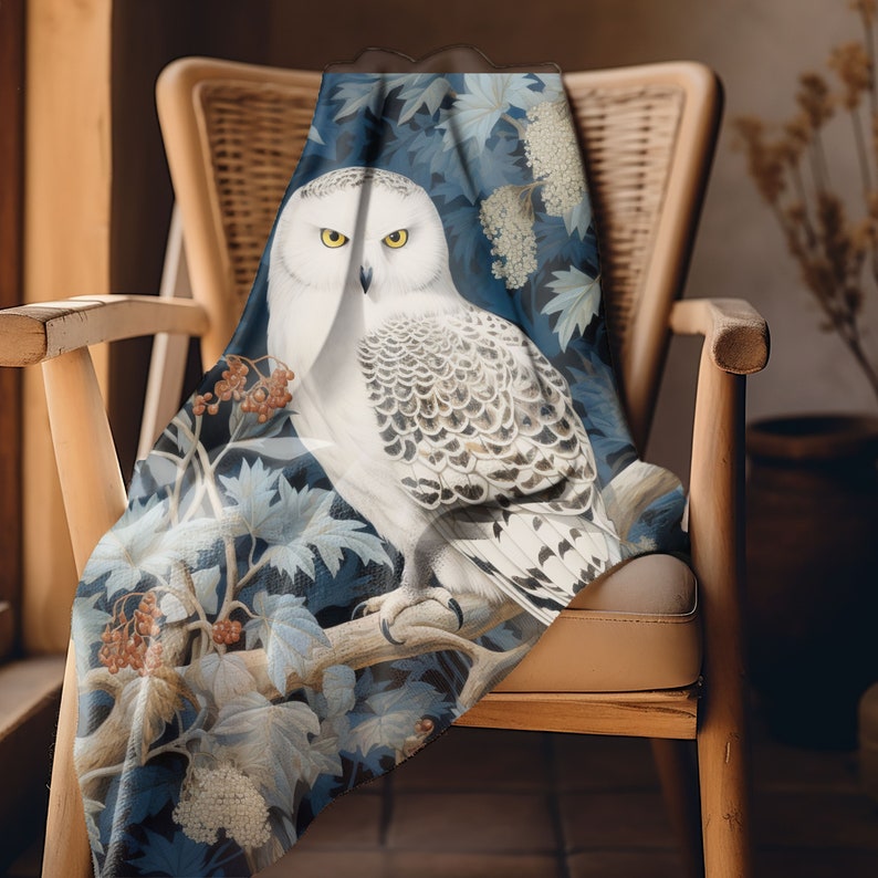 May include: A blue and white throw blanket with a snowy owl perched on a branch. The owl has white feathers with black markings and yellow eyes. The background features a floral design with white flowers and green leaves.