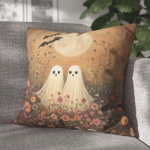 May include: Square decorative pillow featuring a whimsical Halloween design. Two friendly ghost figures stand in a field of flowers under a full moon, with bats flying overhead. The color palette includes warm tones of orange, brown, and cream.