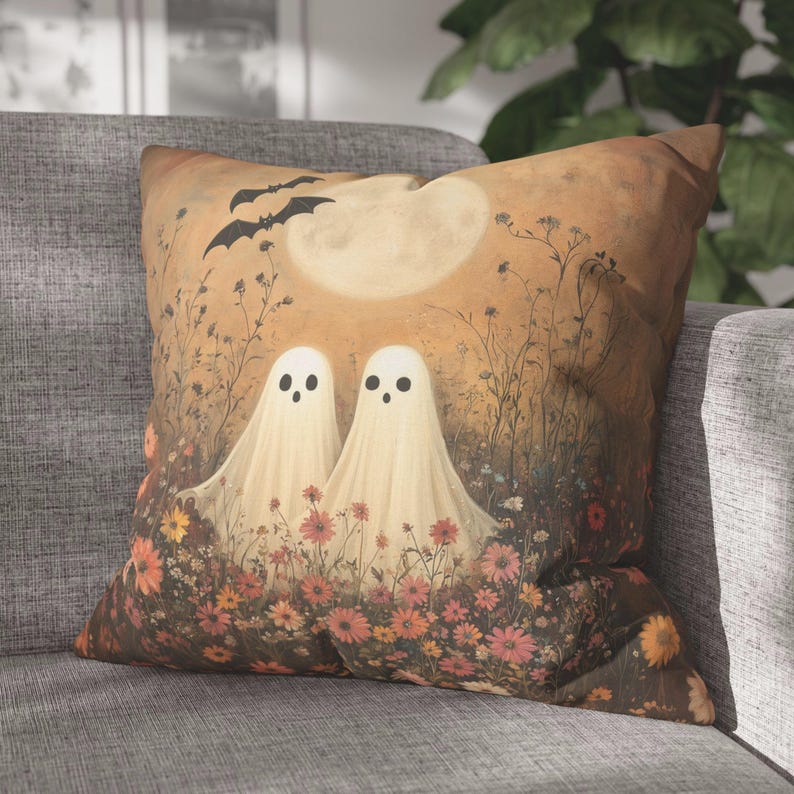 May include: Square decorative pillow featuring a whimsical Halloween design. Two friendly ghost figures stand in a field of flowers under a full moon, with bats flying overhead. The color palette includes warm tones of orange, brown, and cream.