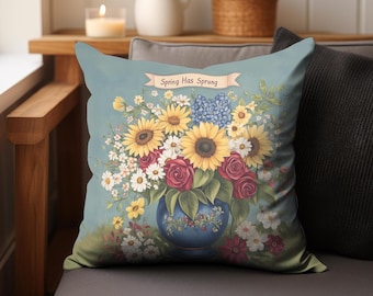 May include: A blue pillow with a floral design featuring sunflowers, roses, and other flowers in a blue vase. The pillow has the text "Spring Has Sprung" on a banner.