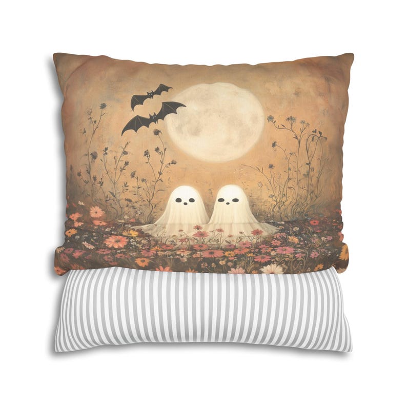 May include: Decorative pillow featuring a whimsical Halloween scene. Two friendly ghost figures sit in a field of flowers under a full moon, with bats flying above. The bottom pillow has gray and white stripes.