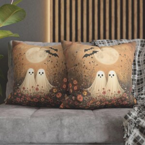 May include: Two decorative throw pillows with a Halloween theme. Each pillow features a whimsical illustration of two friendly ghosts in a field of flowers under a full moon and bats. The pillows have a warm color palette.