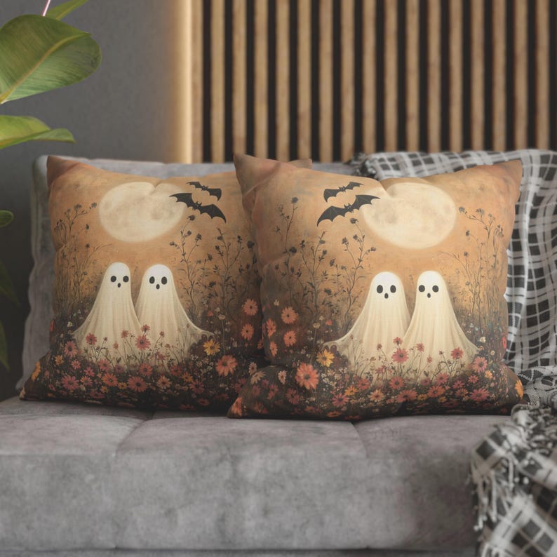 May include: Two decorative throw pillows with a Halloween theme. Each pillow features a whimsical illustration of two friendly ghosts in a field of flowers under a full moon and bats. The pillows have a warm color palette.