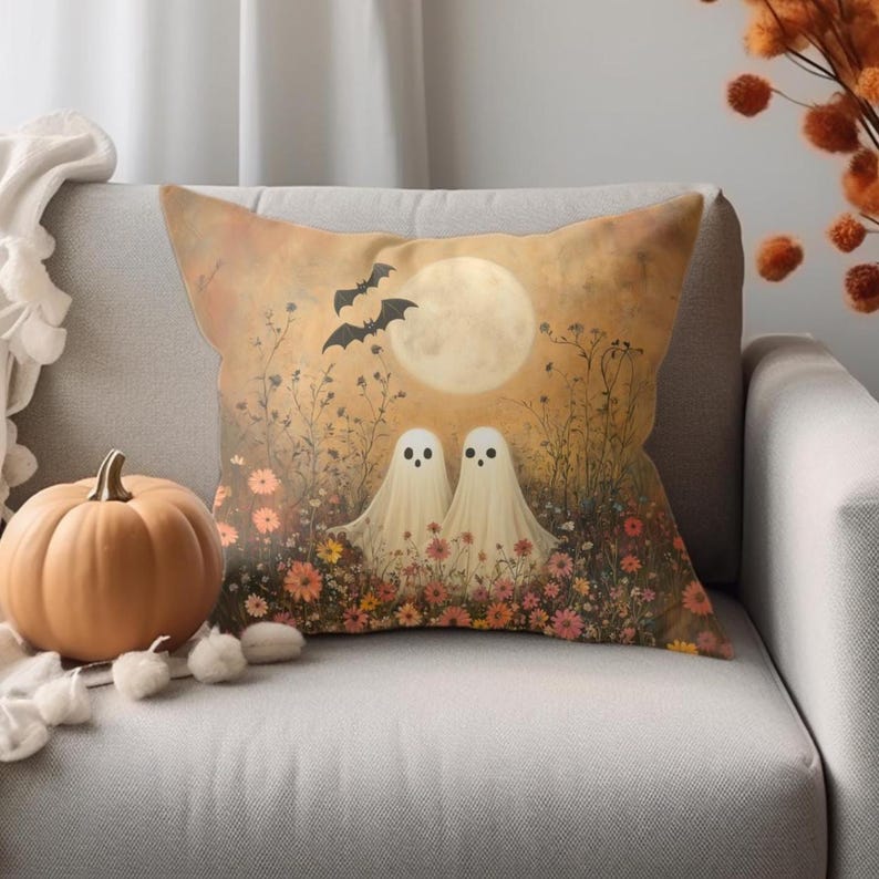 May include: A decorative Halloween-themed pillow featuring two friendly ghosts in a field of flowers under a full moon. Bats fly in the sky above the ghosts. A pumpkin and decorative cotton balls are next to the pillow.