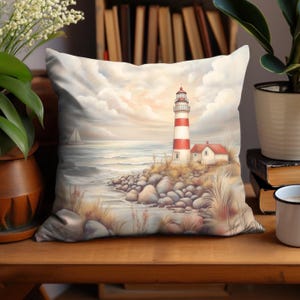 May include: A decorative pillow featuring a coastal scene with a lighthouse. The pillow has a white and red striped lighthouse, a small house, and a sailboat on the ocean. The background has a cloudy sky and the pillow is on a wooden surface.
