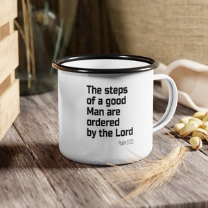 Op de afbeelding: Witte emaille mok met een zwarte rand en handvat. De mok heeft een zwarte tekstcitaat dat luidt: "De stappen van een goede man worden door de Heer geordend" met de verwijzing "Psalm 37:23" eronder.