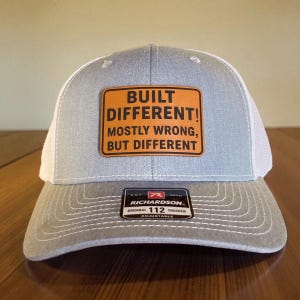 May include: A grey and white trucker cap featuring the text "BUILT DIFFERENT! MOSTLY WRONG, BUT DIFFERENT" on an orange patch. The cap has a curved peak and an adjustable strap.