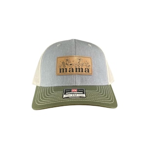 May include: A grey and olive green trucker cap with a beige mesh back. The cap features a brown leather patch with the word "mama" and floral designs. The brim is olive green, and the cap is adjustable.
