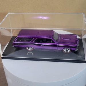 May include: A purple die-cast model of a classic station wagon, displayed in a clear acrylic case. The car features chrome accents, white hood stripe, and red-line tires. The model is presented on a black base.
