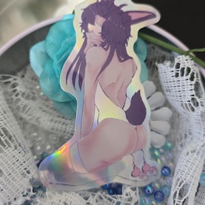 May include: Holographic sticker featuring a seated figure with bunny ears and a tail. The figure has dark hair and is partially nude. The sticker has a rainbow sheen and is placed in a decorative container with blue beads and a flower.