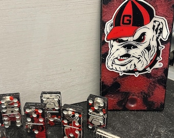 May include: A set of black and red dominoes with a University of Georgia Bulldogs logo. The dominoes are made of resin and have a glossy finish. The dominoes are arranged in a random pattern on a black surface. A red and black sign with the University of Georgia Bulldogs logo is in the background.