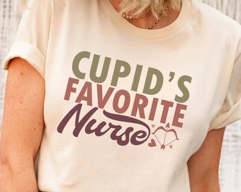 以下が含まれることがあります： ベージュのTシャツ。袖を折り返し、「Cupid's Favorite Nurse」の文字が緑、バーガンディ、紫でプリントされています。テキストの下には弓矢のグラフィックがあります。青いジーンズと組み合わせています。