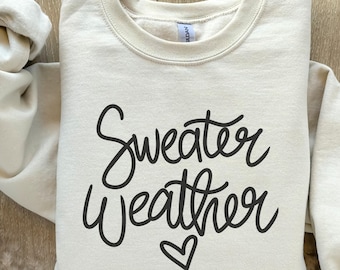 May include: Cream-colored sweatshirt with the words "Sweater Weather" in black script, accompanied by a small heart. The sweatshirt is folded neatly on a wooden surface, showcasing its casual style and cozy appeal.