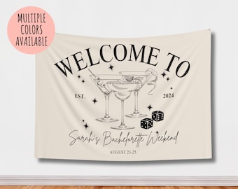 Può includere: Uno striscione in tessuto beige con testo nero che recita "Benvenuti al weekend di addio al nubilato di Sarah, Est. 2024, agosto 23-25". Lo striscione presenta un disegno di tre bicchieri da martini e due dadi.