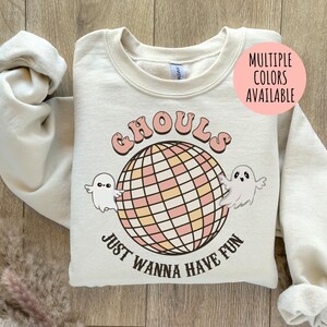 以下が含まれることがあります： レトロなフォントで「Ghouls Just Wanna Have Fun」と書かれたクリーム色のスウェットシャツ。ピンクと黄色の正方形のディスコボールと2匹の漫画の幽霊がデザインされています。「複数の色をご利用いただけます」という言葉がピンク色の円の中にあります。