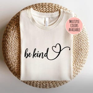 May include: A white t-shirt with the text "be kind" in black cursive lettering and a small heart symbol. The text is centered on the shirt. The shirt is on a woven surface.
