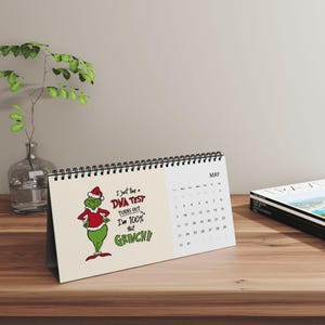 May include: A spiral-bound desk calendar for the month of May featuring a cartoon illustration of the Grinch wearing a red Santa suit. The text reads, I just took a DNA TEST turns out I'm 100% that GRINCH!! The calendar has a cream background.