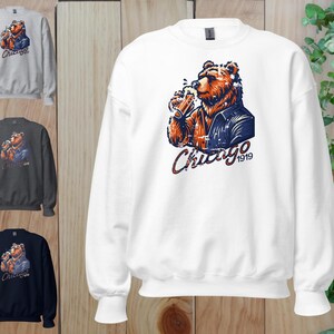 May include: White sweatshirt featuring a graphic of a bear wearing a blue jacket and holding a beer. The bear is looking to the right. The text "Chicago 1919" is below the bear.