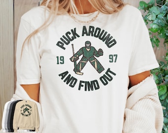 May include: White t-shirt with a green and black graphic print of a hockey player in a goalie stance. The graphic says "Puck Around and Find Out" with the numbers 19 and 97.