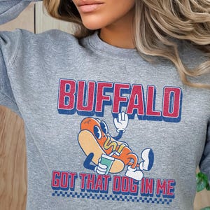May include: A gray sweatshirt with the word "BUFFALO" in red and blue lettering. Below the word is a cartoon hot dog character with the text "GOT THAT DOG IN ME". The sweatshirt has a crew neck and long sleeves.