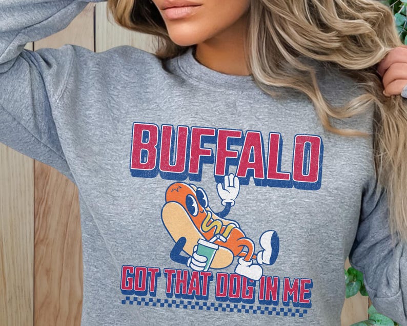 May include: A gray sweatshirt with the word "BUFFALO" in red and blue lettering. Below the word is a cartoon hot dog character with the text "GOT THAT DOG IN ME". The sweatshirt has a crew neck and long sleeves.