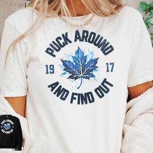 May include: White t-shirt with a blue and white graphic of a maple leaf with the text "Puck Around and Find Out" and the numbers "19" and "17" around the leaf.