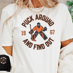 May include: White t-shirt featuring a vintage-style graphic of a hockey goalie in orange and black. The graphic reads "Puck Around and Find Out" with the numbers 19 and 67.
