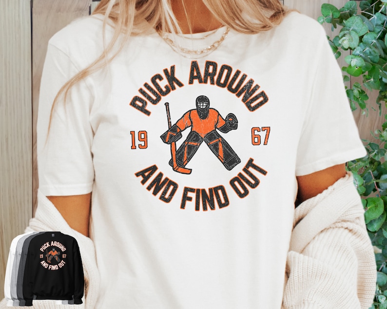 May include: White t-shirt featuring a vintage-style graphic of a hockey goalie in orange and black. The graphic reads "Puck Around and Find Out" with the numbers 19 and 67.
