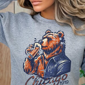 May include: Grey sweatshirt featuring a graphic of a brown bear wearing a blue jacket and drinking from a glass of beer. The text "Chicago 1919" is printed below the bear.