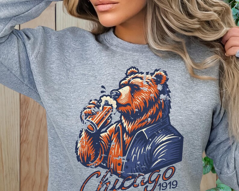 May include: Grey sweatshirt featuring a graphic of a brown bear wearing a blue jacket and drinking from a glass of beer. The text "Chicago 1919" is printed below the bear.