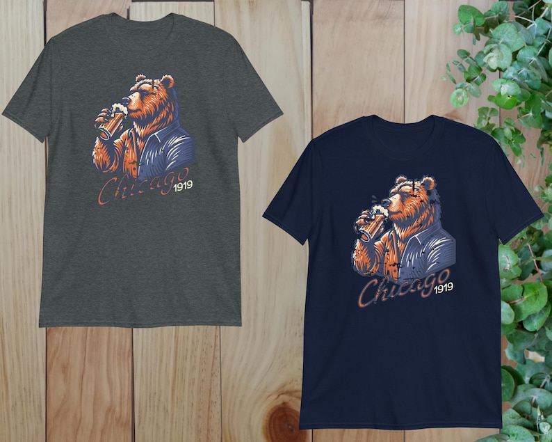 May include: Two t-shirts featuring a graphic of a brown bear wearing a blue jacket and holding a beer. The text "Chicago 1919" is printed on the shirts.