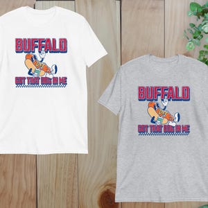 May include: White and grey t-shirts with a graphic design. The design features the word "BUFFALO" in red and blue, above a cartoon hot dog character holding a drink. Below the hot dog is the text "GOT THAT DOG IN ME".
