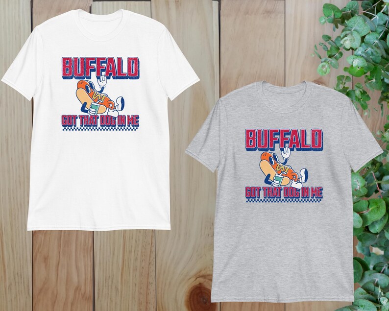 May include: White and grey t-shirts with a graphic design. The design features the word "BUFFALO" in red and blue, above a cartoon hot dog character holding a drink. Below the hot dog is the text "GOT THAT DOG IN ME".