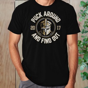 Op de afbeelding: Zwart T-shirt met een gouden afbeelding van een ridderhelm in een cirkel met de tekst "Puck Around 2017 And Find Out".
