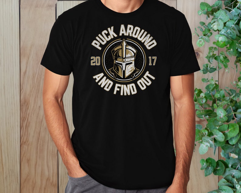 Op de afbeelding: Zwart T-shirt met een gouden afbeelding van een ridderhelm in een cirkel met de tekst "Puck Around 2017 And Find Out".