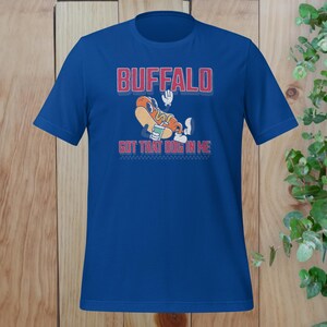 May include: A royal blue t-shirt featuring the word "BUFFALO" in red block letters with a white outline. Below the word is a cartoon hot dog character with the text "GOT THAT DOG IN ME". The shirt has a crew neck and short sleeves.