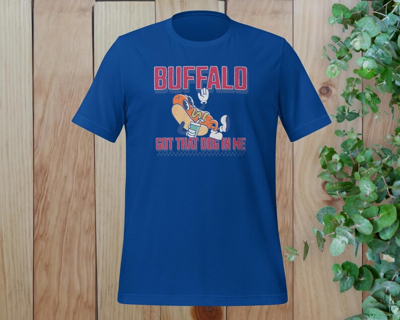 May include: A royal blue t-shirt featuring the word "BUFFALO" in red block letters with a white outline. Below the word is a cartoon hot dog character with the text "GOT THAT DOG IN ME". The shirt has a crew neck and short sleeves.
