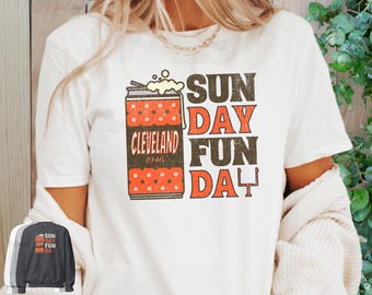 May include: White t-shirt with a distressed graphic featuring a beer can design with the text "Cleveland 1946" and the words "Sunday Fun Day" in orange and brown. The shirt has a vintage, retro style.