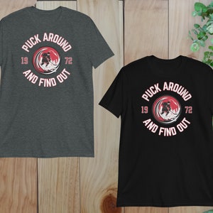 May include: Two t-shirts with a hockey player graphic and the text "Puck Around And Find Out" in a red circle with white text. The numbers 19 and 72 are on either side of the circle. One shirt is dark heather grey and the other is black.